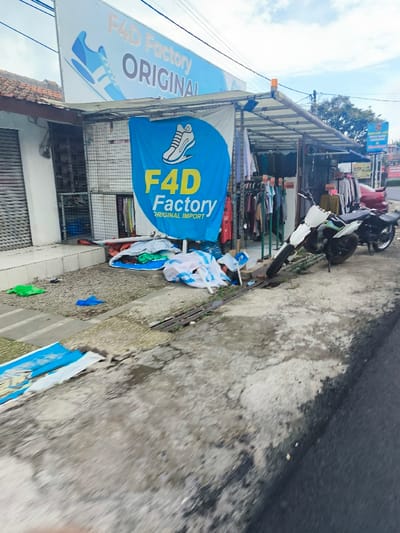 The image shows the exterior of a store called "F4D Factory Original Import", located in Lembang, Indonesia, at latitude -6.822 and longitude 107.61. There is a sign on the storefront with the store name and a blue and yellow logo depicting a sneaker. The sign is above a blue banner displaying the same logo and the words "F4D Factory Original Import". The storefront is mostly obscured by a metal awning covered in a corrugated metal roof. The store has a large window facing the street, and inside it, there are clothes hanging on racks. To the right of the store, there is a white motorcycle parked on the sidewalk. There are several pieces of clothing strewn across the sidewalk in front of the store, along with several blue and white banners. The sidewalk is paved with gray concrete. The street is paved with asphalt. There are no people visible in the image.