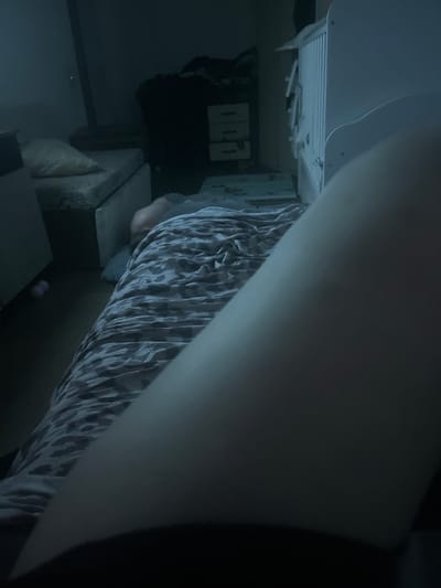 The image depicts an indoor scene, possibly a bedroom or living room, given the presence of what appears to be a bed with patterned bedding and a piece of furniture resembling a headboard or storage unit. A person is partially visible, their legs covered by the patterned fabric, suggesting they are lying down or sitting. The lighting is dim and cool-toned, indicating a low-light environment, perhaps late evening or night. The overall atmosphere is subdued. On the wall behind the bed, there are some objects that look like framed pictures or shelves. To the left, a white ventilation grate is visible, possibly part of an air conditioning or heating system. There are no clear indicators of the weather or a specific time of day beyond the dim lighting. No text is visible in the image. The location context provided is Chelyabinsk, Russia, but there are no specific visual cues within the image to confirm this location.
