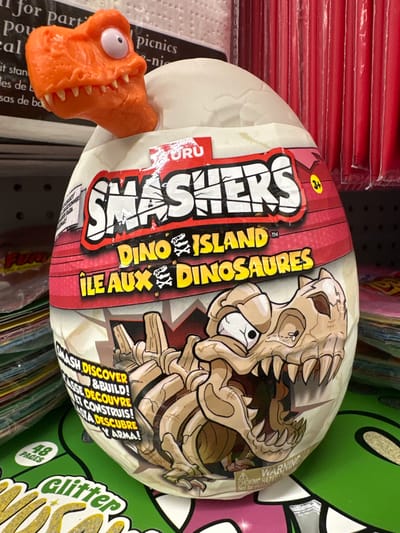 The image shows a "Smashers Dino Island" toy egg with a plastic orange dinosaur head emerging from the top. The egg is predominantly white and covered in colorful graphics featuring cartoon dinosaurs and the "Smashers" logo. Text on the egg includes "DINO ISLAND" and "ILE AUX DINOSAURES". A warning label about choking hazards is visible at the bottom. The egg is placed on a green surface, possibly a shelf in a store, with other products visible in the background, including rolls of red items and stacks of colorful items. The scene appears to be a retail environment. The overall impression is of a toy display in a store.