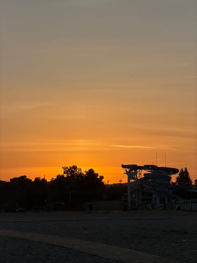 The image displays a coastal landscape captured during either sunrise or sunset, characterized by an intensely orange and red sky that occupies the upper two-thirds of the frame. The sky gradient transitions from a pale, desaturated orange-grey at the zenith, gradually deepening to a vibrant orange and then a fiery orange-red along the horizon. Sparse, wispy horizontal cloud formations are visible in the mid-sky, alongside numerous minute, dark specks identified as birds.
The lower third of the image is dominated by dark, silhouetted elements. The immediate foreground consists of dark-toned beach sand, across which a wooden boardwalk extends diagonally from the lower-left corner towards the center. Above the beach, a dense and irregular line of dark tree foliage forms the terrestrial horizon, obscuring details beyond. To the right of the frame, a large, multi-story water slide structure is prominently silhouetted against the bright sky. This structure comprises a metal framework supporting several intertwining slides, which exhibit dark blue and reddish sections. The appearance of the slides suggests disuse or closure. Faint graffiti is discernible on the lower concrete elements of the slide's support structure. No individuals are present in the scene. This environment is consistent with a beach resort or recreational area located within the city of Catania, Italy.