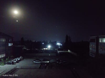 This media file depicts a nighttime scene in Angren, Uzbekistan, likely captured from a high vantage point. The sky is dark and mostly clear, illuminated by a bright full moon and several smaller celestial bodies, possibly stars. Wisps of clouds are visible around the moon, adding a slight atmospheric haze.
The scene includes several buildings, appearing to be apartment complexes or educational facilities, with lights visible in some windows. In the foreground and mid-ground, a parking lot contains several cars. Some of these cars are parked neatly in designated spots, while others are more scattered. There is also a fenced area and what appears to be a covered outdoor seating area or market stall with a few figures present, suggesting some activity or gathering despite the late hour. The general atmosphere is quiet and still, characteristic of a nighttime setting in a residential or semi-urban area.
Notable details include the presence of streetlights and building lights that cast a glow on the surroundings, contrasting with the darkness. The overall lighting is dim, with the moon being the primary natural light source. The image captures the quiet ambiance of a residential area at night, with the moon dominating the sky.
The text "nubia Neo 3 5G" is visible in the bottom left corner, indicating the device used to capture the image.