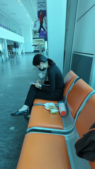 The image captures a woman resting in an airport waiting area, characterized by rows of connected orange cushioned seats with metal frames. The woman, with dark hair tied back, wears a black long-sleeved top, black trousers, and black flat shoes. An eye mask or sleep mask is pulled down over her eyes, suggesting she is trying to sleep. She is seated, leaning forward with her head down, and appears to be holding a purple rectangular object, possibly a phone or small tablet. A silver-colored watch is visible on her left wrist.

On the empty seat next to her, a white thermos with an orange top, some snacks in plastic wrapping, and a white tissue are placed. The floor is a light grey. In the background, a large, modern indoor space with high ceilings and cool blue-white lighting is visible. Architectural features include white walls and ceiling, with some white X-shaped structural supports. A vertical banner advertisement on a wall features a man in a red shirt and sunglasses, holding a bag. Text on the banner reads: "@eraman Shop Duty Free®". The scene depicts a common activity in an airport terminal, likely in Sepang, Malaysia, where travelers rest between flights or during waits.