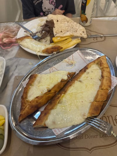 The image depicts two plates of Turkish food, likely in a restaurant setting in Esenyurt, Turkey. The focus is on the dishes: one plate features a cheese-filled pide (a Turkish flatbread) cut into slices and served on a metal platter, with a fork resting on top. The other plate contains what appears to be a döner kebab with fries, wrapped in flatbread. A can of fruit juice is visible near the döner plate. A person wearing a black shirt with a Hello Kitty design is partially visible near the döner plate. The plates rest on a table with printed advertisements. Part of a white dish and a container with pickled vegetables are at the bottom of the frame.