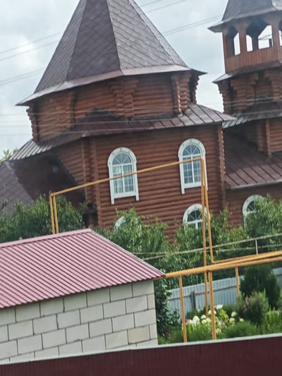 The image captures a close-up view of a wooden structure, possibly a building or a church, with a distinctive multi-faceted dark brown roof. The structure is made of horizontally laid logs, visible in a rich brown hue. Several arched windows with white frames are present on the facade. To the right of the main structure, a smaller tower-like element, also made of wood, rises, featuring open sections. In the foreground, a light-colored brick structure with a red corrugated roof is visible. A yellow metal frame structure runs across the midground, supporting some pipes or conduits. Beyond this, there is lush green foliage, a metal fence, and a garden with white and pink flowers. The sky above is overcast with some visible power lines. The scene appears to be located in Novye Vyselki, Russia, suggesting a rural or suburban setting given the architectural style and surrounding greenery. There are no people or animals visible in the frame.