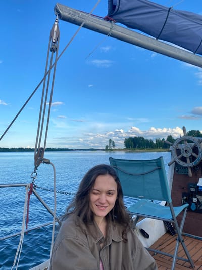 A woman with long, dark, wavy hair winks at the camera while sitting on a sailboat on a body of water, likely a river or lake. She is wearing a light brown jacket over a pink shirt. The sailboat has a large sail deployed and is equipped with a ship's wheel, various ropes, and a blue folding chair. In the background, a tree-lined shore is visible, with some buildings and clouds in the sky. The overall scene suggests a leisurely day on the water.