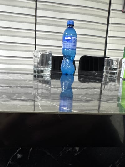 An indoor scene is depicted within the city of Vokil, Bulgaria. A dark gray/black marbled tabletop dominates the foreground, exhibiting a polished, reflective surface. On the tabletop, positioned left to right, are: one clear, empty drinking glass with a thick base, marked with faint 'G' or '2006' lettering; one blue plastic Fanta bottle, partially filled with light blue liquid, exhibiting condensation, with its label facing the viewer; and one clear, empty drinking glass with a thick base, bearing 'G Grann' lettering. All three objects cast distinct reflections on the table surface. The dark, upholstered backs of two chairs are partially visible behind the table; one is behind the left drinking glass, and the other is behind the Fanta bottle and right drinking glass. A portion of a green object is visible on the far right edge of the tabletop. The background features horizontal blinds with alternating light gray and white stripes, bisected by a thin vertical cord on the left. No individuals are present in the frame.