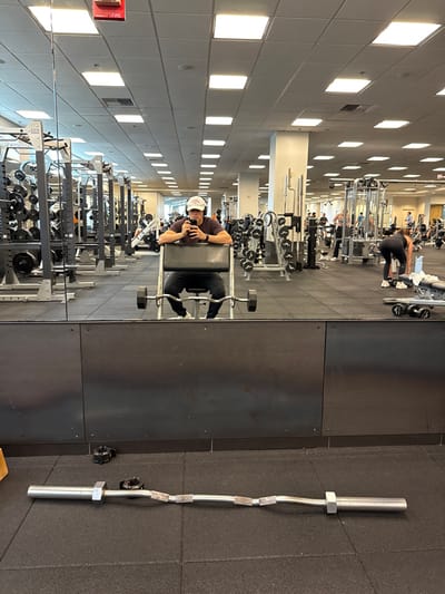 The image shows the interior of a large, well-lit fitness center or gym, located in Boston, United States. The primary subject is a male individual, visible as a reflection in a prominent wall-mounted mirror, seated on a black preacher curl bench. He wears a dark short-sleeved shirt, dark pants, white athletic shoes, black over-ear headphones, and a white baseball cap featuring a red logo (consistent with Callaway Golf branding). He holds a black smartphone in both hands, angled as if capturing a mirror selfie.
In the foreground, on the black rubber mat flooring, lies an EZ curl bar with two black barbell collars positioned nearby. The gym's ceiling is light-colored and features numerous rectangular fluorescent light panels, with a red fire alarm box mounted overhead. The floor throughout the facility is uniformly covered with black rubber mats.
The mirrored reflection reveals an expansive gym space equipped with various weightlifting and exercise machines, including multiple dumbbell racks, power cages, cable machines, and other resistance training apparatus. Several other individuals are visible in the background, exercising. Specifically, a woman in dark athletic wear is observed bending over near a weight bench on the right side of the reflection, appearing to interact with dumbbells or adjust equipment. The environment is modern, organized, and typical of a commercial fitness establishment.