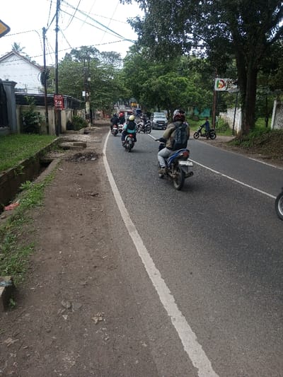 The image depicts an outdoor street scene in Chhenangri_Rishor, Bhutan, under an overcast sky, captured from a low vantage point looking down a paved road that curves gently to the right.
On the asphalt road, a column of multiple motorcycles travels away from the viewer. The nearest motorcycle has a blue body and a dark seat, with a partially visible rear license plate reading "F 4346 UP". Its operator wears a khaki-colored jacket, light-colored pants, and a dark helmet, with a dark backpack. Immediately ahead, another motorcyclist wears a light blue jacket, light pants, and a yellow helmet. Further down the road, at least three additional motorcycles are visible, each with a single rider wearing dark attire and a helmet. Behind the motorcycles, a silver-colored van or SUV is also traveling in the same direction. On the right shoulder of the road, a person in a blue top is seated on a small, dark motorcycle or scooter, facing the camera.
The left side of the road features a dirt shoulder bordering an open concrete drainage ditch. Beyond the ditch, sections of a broken concrete sidewalk are visible. A red rectangular sign displaying "5GB" in white text is affixed to a utility pole. Numerous dark utility poles with a dense network of overhead wires run along this side. Behind the poles, a white building with a dark roof and a white, ornate fence structure is present, accompanied by green trees.
The right side of the road has a dirt shoulder. A large tree with thick green foliage stands prominently. Behind the tree, a white wall with dark discolorations is visible. A pole supporting a colorful sign with red, green, and white elements is located near the roadside. Additional green trees and dense foliage extend into the background on this side.