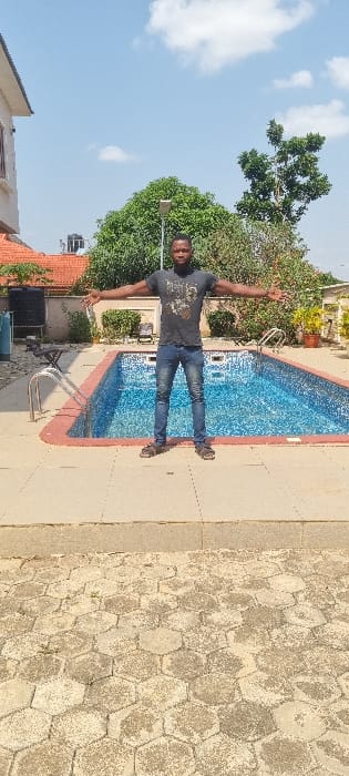 An outdoor scene in Alagbado, Nigeria, depicts a single male subject standing centered in front of a rectangular swimming pool under a partially cloudy blue sky.

The male, appearing to be in his 20s or 30s, possesses a dark complexion and a short beard. He is dressed in a dark grey t-shirt with a light-colored graphic design on the chest, blue denim jeans, and dark sandals. His arms are outstretched horizontally to his sides, and he is looking directly at the camera with a neutral expression.

Behind the subject is a light blue tiled swimming pool with a red coping surround. Two stainless steel handrails with steps are visible on the left side of the pool. The pool is filled with clear, blue-tinted water. Surrounding the pool is a light-colored paved area, primarily consisting of large square tiles directly around the pool and hexagonal pavers in the foreground where the subject stands.

In the background, on the left, is a two-story residential building with a light-colored facade and a dark brown or red sloped roof. A black water storage tank is visible on the roof. Directly behind the pool is a low wall, beyond which are various green shrubs and trees. A pole-mounted solar street light is visible above the wall and between trees. On the right side of the frame, parts of another building with a reddish roof are visible, along with additional vegetation and a potted plant. The sky overhead is clear blue with scattered white cumulus clouds.