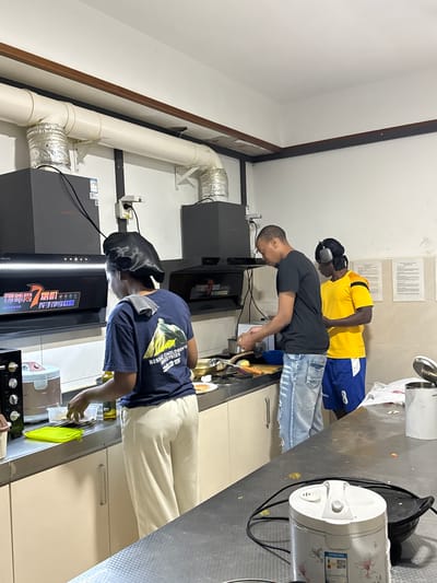 Three young men, appearing to be in their late teens or early twenties, are gathered in a kitchen area, possibly a shared living space or a dormitory kitchen, located in Daxing District, China. The setting is functional, with stainless steel countertops, cabinets, and kitchen appliances like a range hood and a rice cooker visible. The lighting suggests it might be evening or a dimly lit indoor space.

The central figure, wearing a black t-shirt and blue jeans, is actively engaged in cooking, leaning over a cutting board and holding what appears to be a utensil or a bowl. To his left, another young man, dressed in a yellow t-shirt and blue shorts, also seems to be participating in the cooking activity, wearing headphones. The third person, to the left of the frame, is wearing a dark blue t-shirt with text and graphics, and a black head covering, also bent over the counter, possibly preparing food or observing.

Visible text on the blue t-shirt of the person on the far left reads "2025" and "BUSMIC CHILDREN'S INDUSTRIES". The range hood above this individual has Chinese characters printed on it, which likely indicate the brand or model.

The overall atmosphere appears casual and collaborative, suggesting a group of friends or roommates cooking together. There are no strong emotional cues, but the focused postures of the individuals suggest they are concentrating on their tasks. The presence of ventilation pipes on the ceiling hints at the kitchen's functionality for cooking.