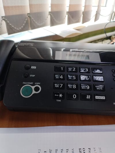 The image shows a close-up view of a black Panasonic fax machine, model KX-FT982, resting on a wooden surface. The fax machine is situated in an indoor environment, likely an office or home, with vertical blinds in the background. The control panel of the fax machine is visible, featuring buttons for various functions such as AOH, STOP, FAX/START, COPY, a dial pad with numbers 0-9 and symbols like asterisk and hash, and function buttons labeled "MONITOR," "HANDSET MUTE," "FL," and others with Cyrillic text. The overall condition of the fax machine appears to be used but functional. No people are present in the image. The location is not specified in the image, but if we assume it to be in Вароніцкі сельскі Савет, Belarus, then the scene depicts a piece of office equipment within that region.