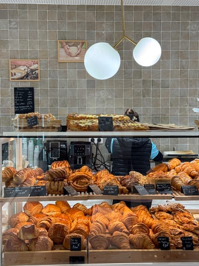 The scene depicts the interior of a bakery or pâtisserie in Asnières-sur-Seine, France. A display counter is filled with a variety of freshly baked goods.
The background wall features light grey-brown square tiles. Two framed pictures adorn the wall, each showing hands working with dough, reinforcing the bakery theme. A modern light fixture with a gold-toned branched frame holds two large spherical white glass globes, providing illumination.
On the upper counter level, two large rectangular savory tarts are visible, one with a small chalkboard label indicating "Focaccia TRom 4,80 €". A wicker basket holds smaller pastries to the left. A black menu board lists various coffee items and prices in French, such as "Café Expresso 1,90 €" and "Chocolat Chaud 3,50 €".
Below the upper counter, behind a glass display, numerous viennoiseries and pastries are arranged on multiple wooden shelves. These include croissants, pains au chocolat, pains aux raisins, and other spiral and layered pastries. Many items have small black chalkboard labels indicating their names and prices in Euros (e.g., "Croissant 1,30 €", "Pain au Chocolat 1,40 €", "Torsade 2,30 €").
Behind the display case, a person is partially visible, facing away from the camera. They wear a dark blue quilted vest over a lighter blue long-sleeved shirt. Their dark hair is pulled back with a light-colored clip. Two black coffee machines (Delonghi brand) and stacks of disposable cups are situated on the counter behind the display, along with electrical outlets and wires. A digital scale is also present on the counter to the right.