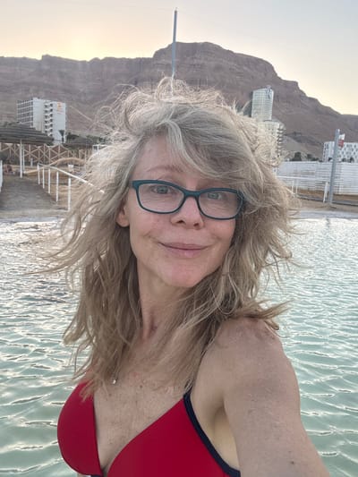This is a selfie taken by a woman with blonde, wind-swept hair, wearing glasses and a red bikini top. She is in the water, which appears to be the Dead Sea, judging by the rippling, mineral-rich surface. In the background, mountains rise up, and there are buildings, including a hotel, suggesting a resort or spa setting in the Tamar Regional Council area of Israel. The sky is overcast or hazy, indicating it might be early morning or late afternoon. The woman appears relaxed and happy.