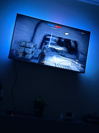 The image shows a television screen displaying a video game. The game appears to be Star Wars: Battlefront II, judging by the character's lightsaber and the game's user interface. The television is mounted on a wall with blue LED accent lighting behind it. Below the television, on a shelf, are a few objects including a small potted plant and some electronic devices. The overall scene suggests a home entertainment setup. The question asks to include the location in the city of Armavir, Russia. However, there is no information in the image to suggest this location. Therefore, the location cannot be determined from the image.