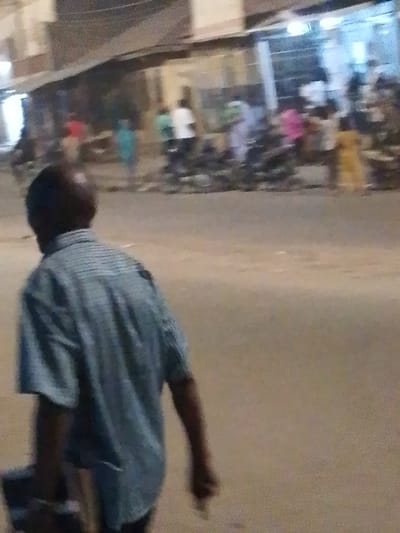 A man walks away from the camera on a street. He wears a blue and white striped short-sleeved shirt and carries a dark object in his left hand. In the background, there is a group of people standing on the street near a building. The scene appears to be at night or in low light conditions. The street is visible with a muted coloration.
