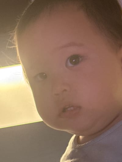 The media file is a close-up, upside-down photograph of a baby's face. The baby appears to be young, with soft skin and large, curious eyes. The lighting is warm and diffused, suggesting an indoor setting, possibly at dawn or dusk. The background is out of focus, but a bright, luminous object or light source is visible, casting a soft glow on the baby's face. The image is composed from a low angle, emphasizing the baby's gaze and creating an intimate feel. The overall mood is one of innocence and wonder. There is no visible text in the image. The location context is Gwangju, South Korea, but this information cannot be discerned from the visual content itself.