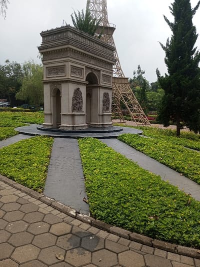 The image depicts an outdoor park setting in Ciherang, Indonesia, featuring miniature architectural replicas under an overcast sky. In the central foreground, a detailed, light-colored replica of the Arc de Triomphe stands on a dark circular base. This structure exhibits classical architectural details, including carved reliefs and multiple arched passages. To the right and behind the Arc de Triomphe, a large, skeletal, light brown lattice structure, representing a section of the Eiffel Tower, is prominently visible. Its lower framework extends into the midground.

The ground around the Arc de Triomphe is landscaped with dark gray paved paths radiating outwards, alternating with dense sections of vibrant green, low-lying ground cover. In the immediate foreground, a pathway constructed from light gray hexagonal pavers runs horizontally across the bottom of the frame, separated from the landscaped area by a concrete curb. Scattered across the scene are various trees: a tall, slender evergreen with dark green foliage is on the far right, and a lighter green weeping tree is visible on the left. The sky is a uniform, light gray, indicating an overcast condition. No individuals or overt actions are present in the scene.