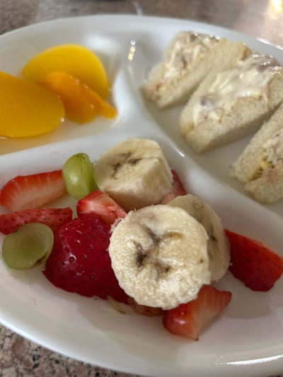 A white, three-sectioned serving plate rests on a light-colored, textured countertop, likely indoors in Thousand Oaks, United States. The top-left section contains several slices of light orange fruit, consistent with canned peaches or apricots, partially submerged in a clear liquid. The top-right section holds three small, triangular sandwiches made with white bread, filled with a creamy, light-colored substance containing red flecks. The bottom section is filled with a medley of fresh fruit, including several circular slices of banana exhibiting minor browning, various cut pieces of red strawberries, and scattered whole or halved green grapes. No people are visible.