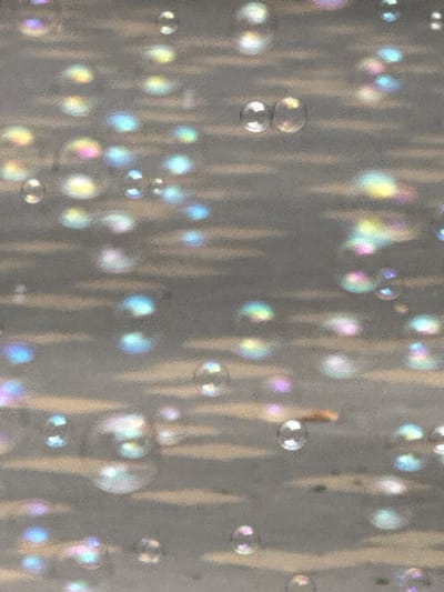 This is a close-up, abstract photograph of soap bubbles floating against a textured, grey background with horizontal stripes. The bubbles are iridescent, reflecting light and colors such as pink, blue, yellow, and white. The lighting suggests an outdoor setting, possibly with sunlight filtering through an overhead structure or canopy, creating the striped effect on the background. There are no people or identifiable landmarks visible, and therefore no specific location in Vinnytsia, Ukraine can be determined from the image. The scene is static and depicts a moment in time with the bubbles suspended in air.