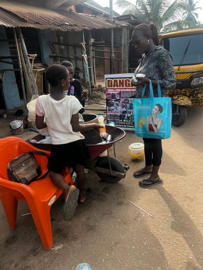 This medium shot captures a lively street scene in Obosi, Nigeria, under bright daylight. In the foreground, a woman stands, looking down at something in her hands. She wears a floral-patterned top and black leggings, and carries a prominent blue reusable shopping bag adorned with an image of a woman wearing jewelry and text such as "Owning gold Having happiness" and "LOVE SOMETHING FOR A LIFETIME." Her feet, in black flip-flops, show pink polished toenails.
To her right, another woman is seated casually on an orange plastic chair, holding a white bottle containing an orange liquid. A brown and black sling bag rests on the chair beside her, which also bears the brand name "Portune". In the background, a yellow commercial tricycle, commonly known as a keke napep, is partially visible, displaying "TVS" and "KING" branding.
Between the two women, a vertical banner advertises "DANG PHONES & PHONE ACCESSORIES SHOP," listing services like "PHONE REPAIR" and stating, "We Specialize On" with further, partially legible text, and "Satisfaction Is Our Priority." A contact number, "TEL 07089232013," is clearly visible. The surrounding environment features buildings with corrugated metal roofing and walls, typical of a bustling local commercial area. Scattered on the ground are white plastic containers, a red metal cart, and a light pink straw. Palm trees are visible in the upper left, adding to the tropical setting. The scene portrays everyday life and commerce in the area.