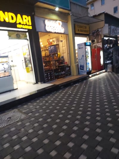 This image presents an angled, slightly downward view of a pedestrian walkway bordered by commercial establishments in an urban environment, likely during the evening in Tiranë, Albania. The pavement is composed of dark grey interlocking blocks with interspersed lighter squares, creating a distinct woven or checkered pattern. A circular metal manhole cover is visible on this surface.
On the left side, several brightly illuminated storefronts indicate active businesses. A prominent sign in the foreground features yellow text on a dark background, partially reading "DARI STOP." Adjacent to it, another shop, possibly a café or convenience store, has a sign that seems to say "Sweet Dream Cafe," featuring a graphic that might be a coffee cup or heart. Its large glass window displays shelves filled with various products. Within this window, a grid-like display board shows numerous small items, along with visible text that includes "Rami çaji kafe" and the number "800."
Further along the street, another business displays a yellow sign with "TOBACCO & PAYMENT" in clear black letters. Outside one of these shops, a red commercial refrigerator stands next to a tall white cooler branded "Medi," which contains bottled water. The scene portrays a lively commercial street. There are no individuals clearly visible, but the well-lit shops suggest ongoing business operations. The overall setting is characteristic of a city street, with multi-story buildings forming the backdrop.