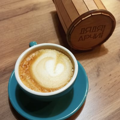 A close-up, high-angle shot shows a cup of coffee with latte art in the shape of a heart, placed on a blue saucer. The coffee cup and saucer sit on a wooden table. To the right and slightly behind the cup is a small, cylindrical wooden container with lettering engraved on it that appears to read "ДЯДЯ АРЧИ". The scene is indoors, likely in a cafe.