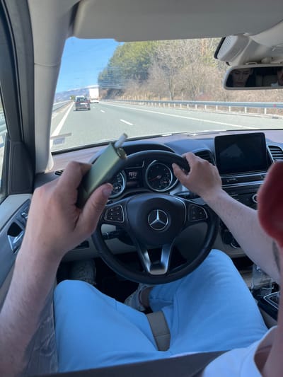 The scene depicts the interior perspective of a vehicle in motion on a multi-lane highway, described as being in the vicinity of Старо село, Bulgaria. The driver, a male, is visible from the chest down, wearing light blue pants and white athletic shoes. His left hand holds an olive green electronic heating device containing a white cigarette stick. His right hand grips the black, three-spoke Mercedes-Benz steering wheel at the 3 o'clock position. The dashboard shows a speedometer indicating approximately 112 units (likely km/h) and an RPM gauge. A digital infotainment screen is present on the center console. Through the windshield, a clear blue sky is visible above a multi-lane asphalt road with a metal guardrail on the right. Sparse trees line the roadside, and distant mountains, one with visible snow, are in the background. Ahead on the highway, a dark-colored SUV is observed in the middle lane, followed by a white truck in the right lane further down the road. The reflection of a female passenger with dark hair is visible in the rearview mirror, looking downwards. A beige seatbelt crosses the driver's lap.