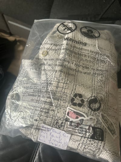 A garment, likely a jacket, is presented inside a clear plastic bag. The bag displays warning symbols and text in multiple languages, including English, Italian, and French, regarding suffocation hazards and proper disposal. There are also recycling symbols and stickers featuring stylized cars on the bag. A handwritten note in German is attached to the bag, expressing thanks for the purchase and well wishes for wearing the garment. The background suggests an indoor setting, possibly a home environment, with a dark, textured surface that appears to be upholstery. The image does not provide any specific information about the city of Gelsenkirchen, Germany.
