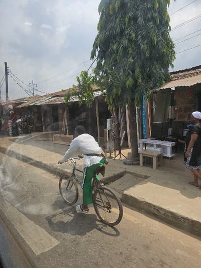 This image captures a daytime street scene in Agege, Nigeria. The primary subject is a male figure, observed from a slightly elevated rear-left perspective, riding a dark-colored bicycle on a paved road. He wears a white long-sleeved top, green trousers, and white slip-on sandals, with a dark item draped over his left shoulder and a white sash around his waist. His shadow is cast on the road surface.

The road features faint white markings and is flanked by concrete sidewalks. A woman in a dark patterned dress and white head covering is partially visible walking on the right sidewalk.

To the right, a row of single-story commercial structures lines the street, constructed with brick and wood, topped with weathered corrugated metal roofs. Various items, including light-colored furniture (tables/benches) and metal bars, are displayed outside these establishments, suggesting furniture or hardware vendors. A tall, slender tree with dense green foliage stands prominently on the right side between the sidewalk and the shops.

On the left side of the street, a wooden utility pole supports multiple overhead wires that crisscross the sky. The sky is partly cloudy, and strong shadows indicate bright daylight conditions. A slight glare is present in the lower-left foreground, suggesting the image was captured through a window.