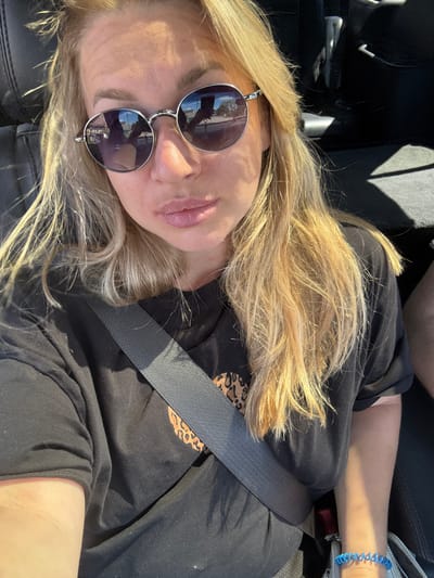 A woman with blonde hair, wearing sunglasses and a black t-shirt, is seen from the chest up, seated in a car. She has on a grey seatbelt and a blue bracelet. The background shows a dark car interior and the sunlight streaming in. The image is a selfie taken inside a vehicle. No specific location in Valday, Russia is identifiable.