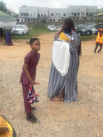 In Galadimawa Village, Nigeria, on a gravelly, dusty ground, a young boy in a maroon traditional outfit stands to the left of the frame, holding a bundle of red, white, and blue fabric in his right hand. He is looking to his left. Behind him, a woman with braided hair wears a long, flowing black and white patterned dress and carries a baby on her back, wrapped in a white cloth. The baby is wearing a yellow shirt. Further in the background, several vehicles are parked, including a white SUV and a dark SUV. A person wearing a bright orange safety vest is visible in the upper right quadrant of the image, walking away from the camera. A building with multiple windows and a flat roof is situated in the background, suggesting an urban or semi-urban setting within the village. A green structure with signage is present on the far left, and a few trees and bushes are scattered throughout the scene. The sky is overcast.