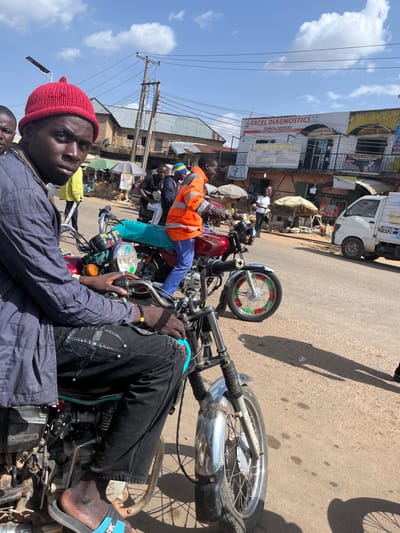 A medium shot captures a man wearing a red knitted beanie and a blue quilted jacket, sitting on a motorcycle in Gwong, Nigeria. He is looking directly at the camera with a serious expression. His black pants are decorated with a criss-cross pattern and silver studs. His right foot is visible, wearing a black and blue sandal. Another motorcycle, red and maroon, is parked next to him, with a turquoise bag on its seat. In the background, several other people are present, some wearing bright orange or yellow jackets, and a white truck is parked on the right side of the street. Buildings with signage, including "EXCEL DIAGNOSTICS," line the street, indicating a commercial area. The scene is set outdoors on a sunny day with a partly cloudy sky.