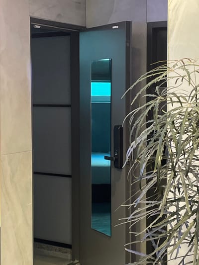 The image depicts an indoor scene, likely a hallway or entryway, in Jongno-gu, South Korea. A modern, dark-colored door, equipped with a black digital lock on its right side, is partially ajar. The door features a central, vertical, reflective panel, which appears to be tinted blue. Through the open space and the reflective panel, a room interior is visible, characterized by a prominent, horizontal strip of bright blue light emanating from an unseen source, possibly a window with horizontal blinds or a light fixture. Below this light, a light-colored surface, consistent with a bed or floor, is faintly illuminated. To the left of the door, a light-colored, possibly textured or paneled, wall is visible. In the right foreground, a large potted plant with numerous long, slender, green leaves partially obscures the view of the door and the wall beyond. No individuals, explicit actions, or direct interactions are visible within the frame.