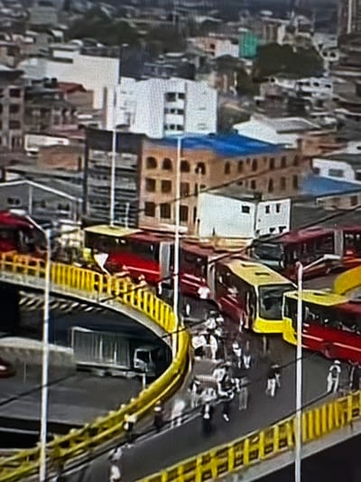The image displays an elevated view of a busy urban transportation hub within Bogota, Colombia, featuring the TransMilenio Bus Rapid Transit system. A prominent, curved pedestrian bridge with bright yellow railings is central to the foreground and mid-ground. Beneath this structure, an elevated roadway hosts multiple red articulated TransMilenio buses, some appearing to be stopped at a station platform. At least two buses with distinct yellow fronts, characteristic of feeder routes, are also visible on the elevated roadway; one is centrally located, facing partially right.
Numerous individuals, identifiable as commuters or pedestrians, are present on the bridge and surrounding elevated platforms. They are actively walking, with no specific interactions or actions of conflict discernible. Their attire is typical urban casual.
The background reveals a dense cityscape comprised of multi-story buildings constructed from various materials and colors. Several structures feature flat roofs, some covered with distinctive blue tarpaulins or roofing materials. Overhead streetlights and utility lines are visible. The scene is illuminated by ambient daylight, suggesting regular operational hours.