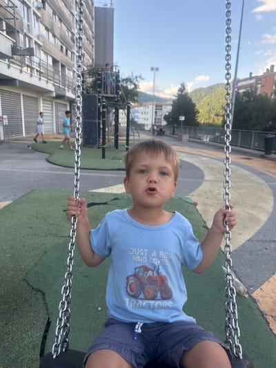 A young Caucasian boy with blond hair and blue eyes is sitting on a swing, holding onto the chains with his hands. He is wearing a light blue t-shirt with a graphic of a red tractor and the text "JUST A BOY WHO LOVES TRACTORS". He is also wearing blue shorts. The background shows a playground with a colorful, curved rubberized surface. In the distance, there are buildings and trees, suggesting an urban or suburban setting. The sun appears to be shining, casting shadows and illuminating the scene. The boy has an expressive look on his face, with his mouth slightly open as if speaking or reacting. The image is oriented vertically, with the top of the boy's head towards the left.