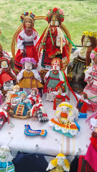 The image shows a collection of handmade dolls displayed on a white, striped cloth. The dolls vary in size and style, with many dressed in traditional Ukrainian folk costumes featuring embroidered patterns and colorful ribbons. Some dolls are adorned with floral wreaths and beaded necklaces. In the background, there's a glimpse of green grass, suggesting an outdoor setting. The dolls are arranged in a somewhat cluttered, artistic manner, some sitting in woven baskets, while others are placed directly on the cloth. In the center of the display, a small, blue embroidered bird figurine is prominently featured. The overall impression is that of a market stall or craft fair. While the image itself does not contain explicit references to Smila, Ukraine, the style of the dolls suggests a possible connection to Ukrainian folk art traditions.
