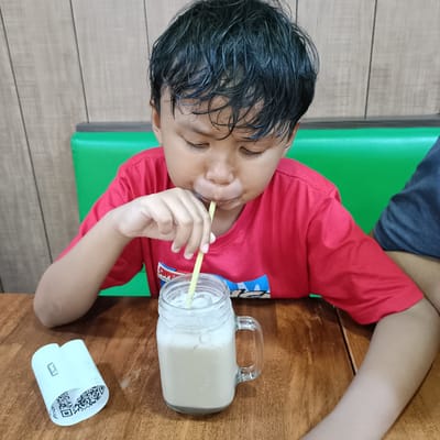 The image shows a young boy in a red t-shirt drinking a milky beverage from a glass mug with a straw. He is sitting at a wooden table, and a receipt with QR codes is visible on the table to his left. In the background, a green booth and a wood-paneled wall can be seen, suggesting an indoor setting such as a restaurant or cafe. The boy's eyes are closed as he sips the drink, and his cheeks are puffed out. Another person, partially visible in a dark blue shirt, is seated to the boy's right. The lighting appears to be artificial, indicating it could be any time of day. The presence of a receipt with QR codes might suggest a recent transaction. The overall mood is relaxed and casual.