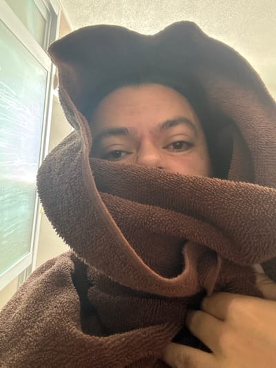 A single individual is depicted in an indoor close-up within the city of San Juan Cuautlancingo, Mexico. The person's face is partially obscured by a brown, terry cloth towel wrapped around their head and lower face. Only the eyes, eyebrows, forehead, and bridge of the nose are clearly visible, with the individual looking directly at the camera. A hand, consistent with the visible skin tone, is present in the lower right corner, appearing to grasp or adjust the towel. The immediate background to the left features a vertical window with a white frame, displaying light reflections and a textured surface on the glass. Above the person's head, a light-colored, textured ceiling is visible.
