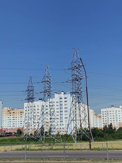 The image shows a clear blue sky above a row of tall, industrial power pylons in the foreground. Behind the pylons, several multi-story apartment buildings are visible, characteristic of urban residential areas. The buildings appear modern, with numerous windows and balconies. Between the pylons and the apartment buildings, there is a strip of green grass and some trees. In the far background, more buildings and vegetation can be seen. In the very bottom of the image, a section of a road and a fence are visible. The scene appears to be a typical urban landscape with infrastructure for electricity transmission running through a residential district.