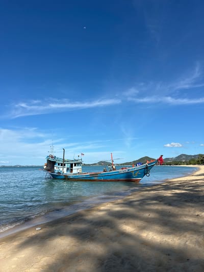 A bright, sunny day unfolds over the ocean and shoreline of Baan Tai, Thailand, with a deep blue sky dotted with wispy white clouds and a sliver of moon visible. A vibrant blue fishing boat, with lighter blue trim and an orange hull, rests partially aground on the sandy beach. The boat displays registration numbers "TS5204H" and other markings in Thai script. A Thai flag flies from its stern. The boat's deck is visible, with some structure and what appears to be netting. The water is calm and shallow near the shore, with gentle waves lapping the sand. The beach is composed of light-colored sand, with scattered shadows suggesting trees nearby. In the background, a lush green coastline with rolling hills stretches across the horizon, lined with palm trees. The scene is devoid of any visible people or animals, presenting a serene, untouched coastal landscape.