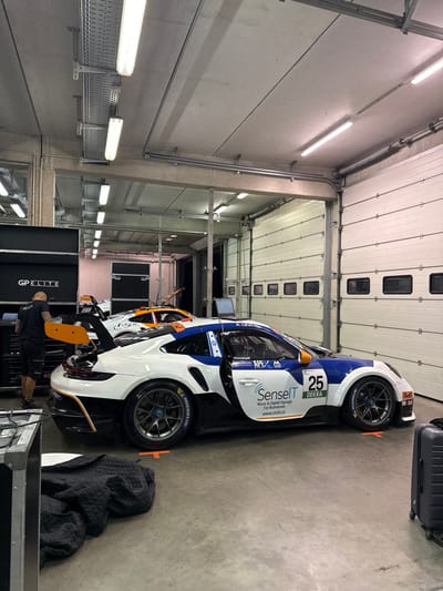 This image depicts an indoor garage or pit box environment, characteristic of a motorsport facility, located in Hohenstein-Ernstthal, Germany, likely at the Sachsenring circuit. The floor is concrete with orange T-shaped markings for vehicle positioning. The ceiling is concrete, equipped with multiple rows of fluorescent tube lights and exposed metal cable trays and conduits. A large white sectional garage door with multiple rectangular windows is closed on the right side of the frame.
Two race cars are present. In the mid-ground, a white, blue, and black Porsche 911 GT3 Cup car, bearing the number 25, is centrally positioned. Its livery includes sponsor logos such as "SenseIT," "DEKRA," "Michelin," and "Mobil," with an Israeli flag decal and the name "A. LEVI" above the driver's side window. The driver's door is open. The car is equipped with dark multi-spoke wheels and Michelin tires. Behind this vehicle, a second race car, predominantly orange and white with a "Corset" logo, has its rear engine cover open, suggesting active maintenance. A laptop computer on a stand is situated between the two cars.
On the left side of the frame, a single male individual is observed from behind. He wears a black t-shirt, black shorts, and black shoes, and has a shaved head. He is standing adjacent to a black "GP ELITE" branded toolbox or equipment cabinet, appearing to be engaged with items on or near it. Several large, dark equipment cases are stacked behind him. In the immediate foreground on the left, a black quilted blanket or cover is crumpled on the floor. A black hard-shell suitcase is partially visible on the far right edge of the frame. The scene indicates a period of vehicle preparation or servicing within a professional motorsport setting.