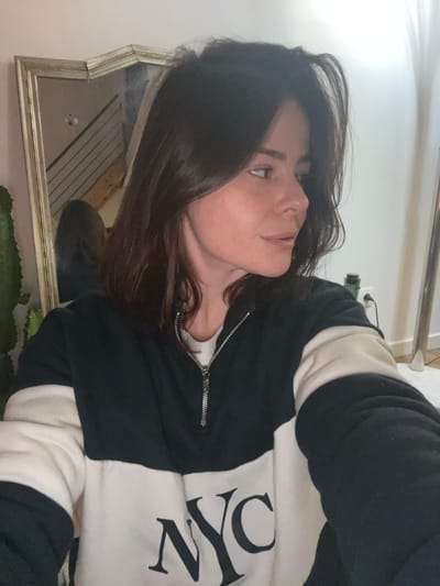 A woman with dark hair looks upward in a selfie. She wears a black and white sweatshirt with "NYC" emblazoned on the front. The garment has a zipper halfway down the chest. The photo is slightly tilted and a gold-framed mirror is visible behind her, reflecting a blurred background. Part of a plant is seen near the mirror. The overall lighting in the photo is soft.