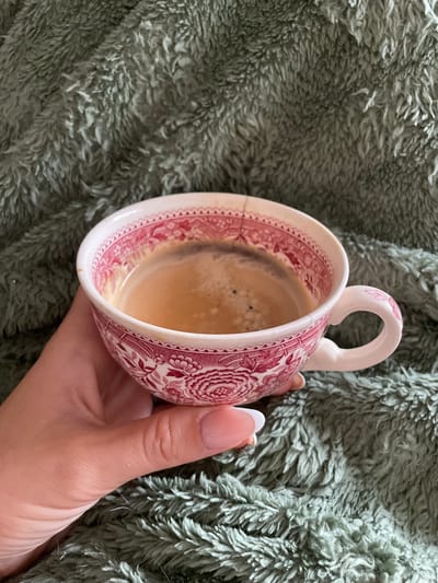 A person's hand holds a vintage coffee cup filled with dark coffee. The cup has a white ceramic base with a detailed red floral pattern around the rim. The coffee has a layer of crema on top. The background is a textured, soft, green blanket. There is no discernible information about the location in Pernik, Bulgaria.