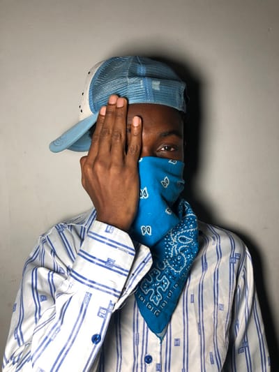 A person wearing a blue bandana over their nose and mouth and a blue trucker hat poses against a gray wall. One hand obscures part of their face. They are wearing a long-sleeved white shirt with blue horizontal stripes and small blue rectangular boxes printed on it. The shirt is buttoned up to the collar. The person is slightly angled.