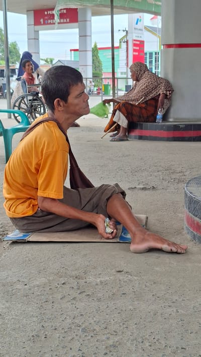 The image depicts a street scene at a gas station in Keudebayu, Indonesia. In the foreground, a man in an orange t-shirt and brown pants sits on a piece of cardboard on the concrete ground. He is holding a few bills in his right hand and is looking to his left. His left leg is extended, and his right leg is bent. A dark brown strap is slung over his left shoulder. In the background, a woman wearing a patterned headscarf and orange and white clothing sits leaning against a pillar. She is holding a plastic bottle of water and a stick. To the left of the foreground man, a person in a wheelchair is visible, with another person wearing a blue head covering standing behind them. The gas station has red and white pillars and signs indicating fuel prices. The ground is a rough concrete surface. The lighting suggests it is daytime.