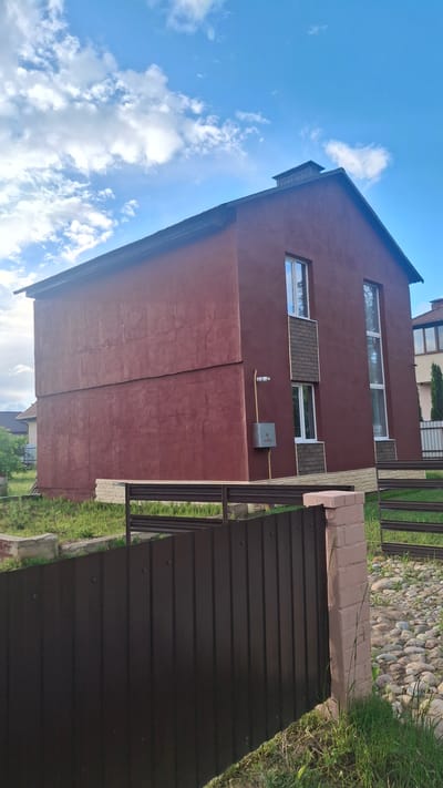 The image shows a two-story, dark red stucco house with a dark brown roof and a prominent chimney. The house has several windows, some of which are tall and narrow, and others are shorter and wider. There are decorative brick accents around some of the windows. A gray electrical box is mounted on the exterior wall, with a yellow conduit running from it. The house is surrounded by a dark brown metal fence in the foreground, with a brick pillar marking one of the fence posts. Beyond the fence, there is a yard with green grass and some scattered stones. In the background, other houses and trees are visible under a bright blue sky with scattered white clouds. The scene appears to be a residential area in Stowbtsy, Belarus, likely a suburban or rural setting given the amount of green space.