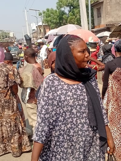 A woman with a dark headscarf and light patterned top is facing left. She is surrounded by other people and various items, likely in a marketplace. Buildings and poles are visible in the background under a bright sky. Another woman wearing a leopard print dress is seen behind the first woman. The scene appears to be outdoors during the daytime.