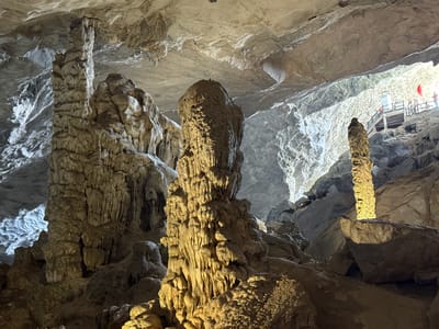 A wide-angle interior shot captures the grandeur of a vast cave system in Ha Long Ward, Vietnam, likely a tourist attraction. The scene is dominated by massive, intricately textured stalagmites and columns, formed from centuries of mineral deposits, rising from the cave floor towards the high, uneven ceiling.
In the foreground and midground, several large rock formations are visible. A particularly prominent, thick column with a rough, layered texture stands centrally, illuminated from below by a warm, yellowish light, highlighting its contours. To its left, another impressive column, appearing more weathered and rugged, extends upwards. On the far right, a slender, light-colored stalagmite also receives focused illumination. The surrounding cave walls and ceiling are a mix of smooth and rippled gray rock, with some areas showing curtain-like stalactite formations.
Towards the upper right of the image, a bright opening or heavily illuminated section of the cave is visible, suggesting an exit or a significant chamber. A wooden viewing platform with railings and stairs is constructed within this brightly lit area, indicating human access and exploration. Several people, appearing small in scale due to the distance, are standing on this platform, observing the cave. A small red flag is visible near the top of the platform, possibly indicating a tour group or a national marker.
The lighting in the cave is a mix of artificial spotlights highlighting specific formations and natural or very strong artificial light emanating from the distant opening, creating dramatic contrasts of light and shadow throughout the cavern. No discernible text is visible in the image.