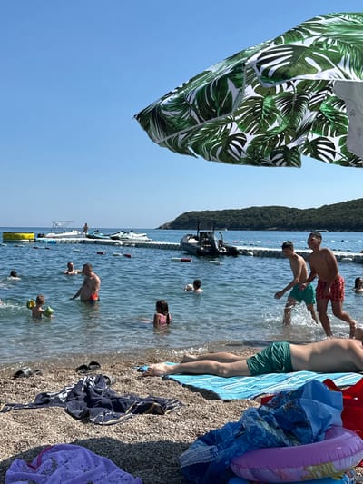 A wide shot captures a sunny beach scene in Prijevor, Montenegro, with a clear blue sky and calm, sparkling water. In the foreground, a pebble beach is dotted with towels and beach accessories. A person lies on a blue striped towel, wearing green swim trunks. Nearby, a dark blue towel is spread out, and a purple towel is partially visible.

The water is populated with several individuals enjoying the ocean. Children wear floatation devices, and adults swim and play. In the middle ground, a white boat and two jet skis are visible near a floating barrier. The background features a lush, green, treed hillside, characteristic of the Montenegrin coast. An umbrella with a tropical palm leaf pattern is partially visible at the top of the frame, adding to the beach ambiance. The overall impression is one of a relaxed summer day at the seaside.