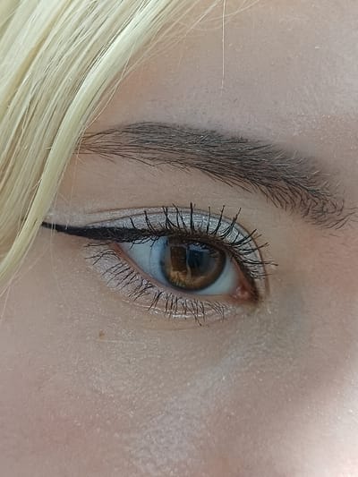 A high-resolution, close-up photograph captures the left eye and surrounding facial skin of an individual, documented in 서대문구, South Korea. The subject presents with light-toned skin, exhibiting visible texture and a singular small blemish inferior to the lower eyelid. Blonde hair is visible in the upper-left quadrant. The eyebrow is dark and groomed. The left eye is open, revealing a brown iris with a rectangular light reflection, and a white sclera. Cosmetic enhancements include light, shimmering eyeshadow on the upper eyelid, a distinct black winged eyeliner applied to the upper lash line, and black mascara darkening and extending both upper and lower eyelashes.